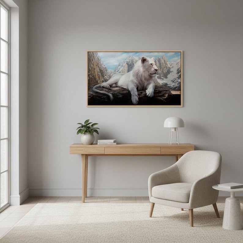 May include: A framed artwork depicts a white lion resting on a log, with a mountain range in the background. The artwork is displayed on a light gray wall above a wooden desk with a small plant, a lamp, and a beige armchair.