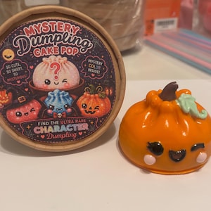 May include: A round cardboard container labeled Mystery Dumpling Cake Pop with colorful graphics of cartoon dumplings and pumpkins, alongside a small, glossy orange pumpkin-shaped dumpling figurine with a brown stem and a smiling face.