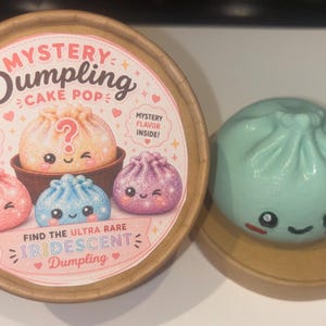 May include: A round cardboard container labeled Mystery Dumpling Cake Pop with illustrations of colorful dumplings and text including Find the Ultra Rare Iridescent Dumpling. Beside it sits a light green dumpling toy with a smiling face.