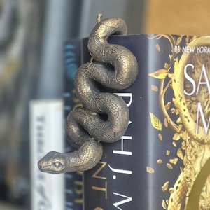Könnte beinhalten: Ein bronzefarbener Schlangen-Lesezeichen, das sich um den Buchrücken windet. Das Lesezeichen hat detaillierte Schuppen und einen realistischen Kopf. Das Buch hat einen schwarzen Einband mit goldenen Buchstaben und Akzenten.