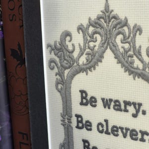 Op de afbeelding: Geborduurde kunst met een grijze sierlijke rand en de tekst "Be wary. Be clever. Be good." De borduursel staat op een witte, getextureerde achtergrond, geplaatst in een donkergrijze passe-partout en lijst.