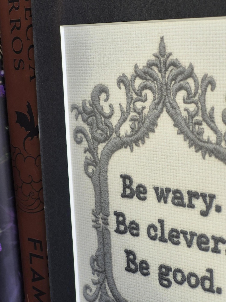 Op de afbeelding: Geborduurde kunst met een grijze sierlijke rand en de tekst "Be wary. Be clever. Be good." De borduursel staat op een witte, getextureerde achtergrond, geplaatst in een donkergrijze passe-partout en lijst.
