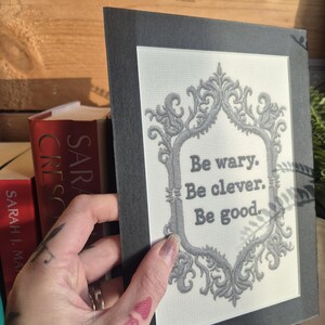 Op de afbeelding: Een ingelijste print met de tekst "Be wary. Be clever. Be good." in een decoratieve grijze lijst. De print wordt voor een houten achtergrond en een stapel boeken gehouden.