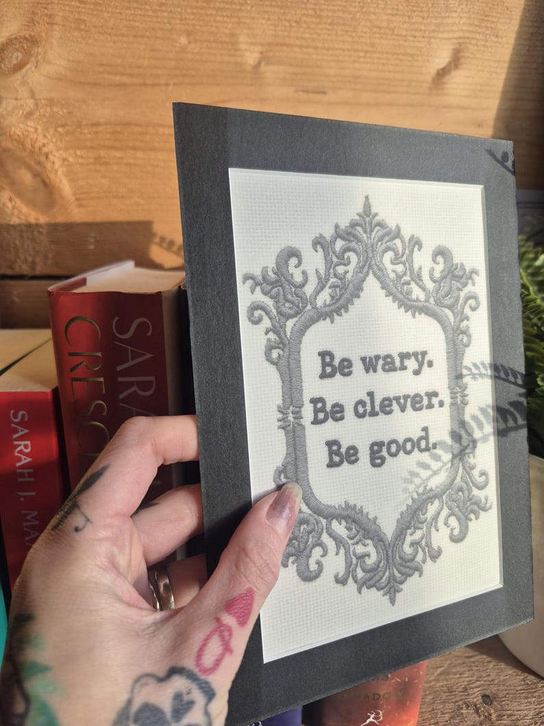 Op de afbeelding: Een ingelijste print met de tekst "Be wary. Be clever. Be good." in een decoratieve grijze lijst. De print wordt voor een houten achtergrond en een stapel boeken gehouden.