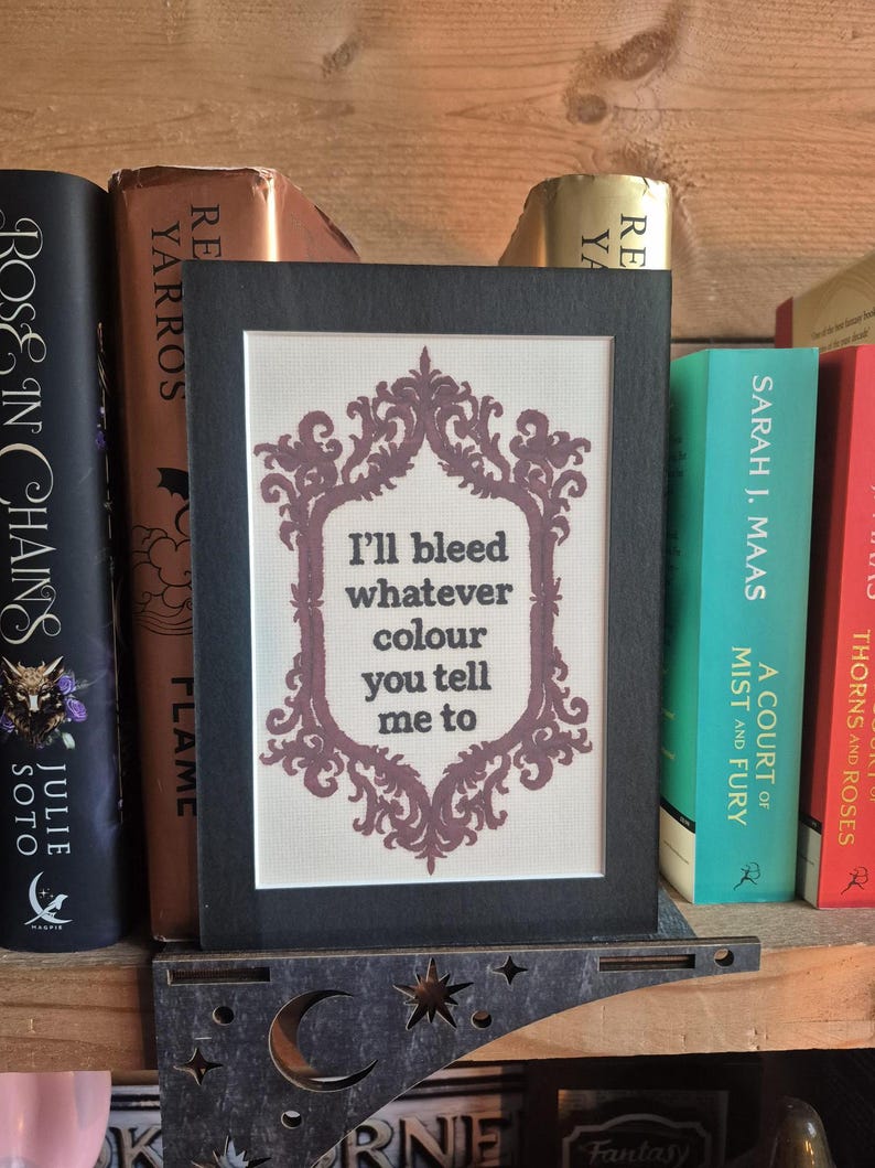 K&ouml;nnte beinhalten: Ein gerahmtes Kreuzstichbild mit dem Zitat "I'll bleed whatever colour you tell me to" in einem dekorativen Rahmen. Der Rahmen ist schwarz und der Text ist dunkelviolett. Die gerahmte Kunst wird auf einem Holzregal mit B&uuml;chern ausgestellt.