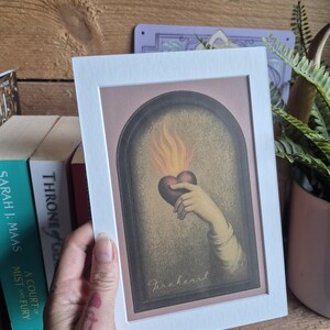 May include: A rectangular art print in a white mat frame featuring an illustration of a hand holding a dark heart with orange flames rising from it, set within an arched border. The word Fireheart is written in cursive script at the bottom.