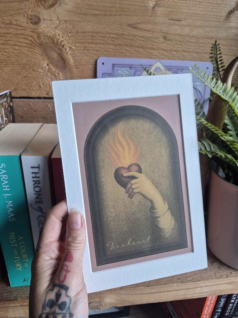 May include: A rectangular art print in a white mat frame featuring an illustration of a hand holding a dark heart with orange flames rising from it, set within an arched border. The word Fireheart is written in cursive script at the bottom.
