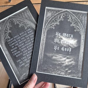 May include: Two framed art prints with gothic arch designs. One print features the text "Be Wary, Be Clever, Be Good" in elegant script. The other print displays a passage of text. Both prints are in black and white.