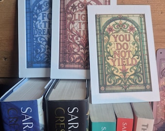 May include: Three framed art prints with inspirational quotes, displayed above a stack of books. The prints feature stained-glass style designs with floral motifs and the phrases "FOR THE DREAM", "LIGHT IT UP", and "YOU DO NOT YIELD". The books are by Sarah J. Maas.
