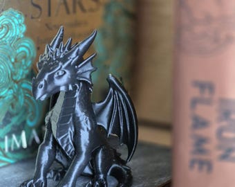 May include: A black dragon figurine with detailed wings, claws, and a spiky head. The dragon is sitting on a gray surface. The background includes a book with the words "FALLEN STARS" and "IRON FLAME".