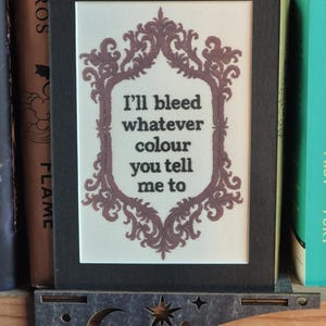 K&ouml;nnte beinhalten: Gerahmte Kreuzstichkunst mit schwarzem Rahmen. Das Kunstwerk zeigt einen dekorativen, mauvefarbenen Rahmen, der den Text "I'll bleed whatever colour you tell me to" umgibt.