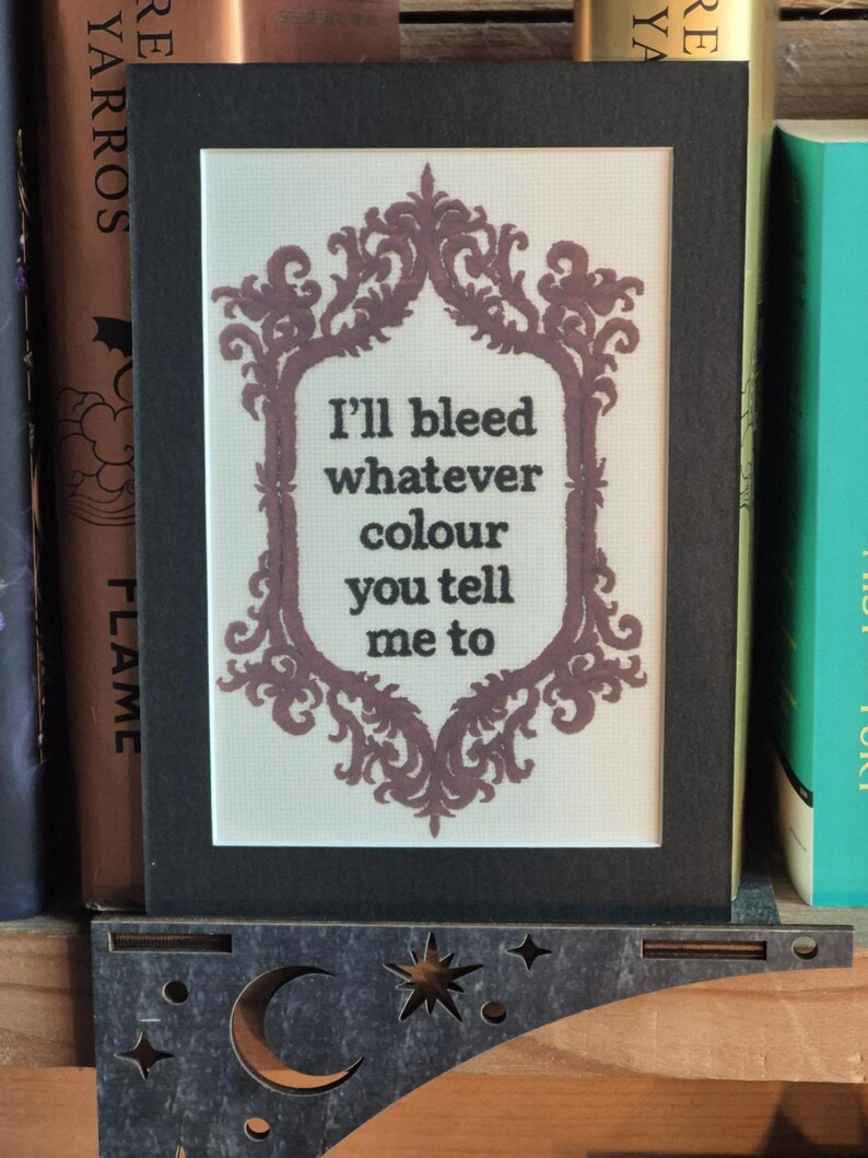 K&ouml;nnte beinhalten: Gerahmte Kreuzstichkunst mit schwarzem Rahmen. Das Kunstwerk zeigt einen dekorativen, mauvefarbenen Rahmen, der den Text "I'll bleed whatever colour you tell me to" umgibt.