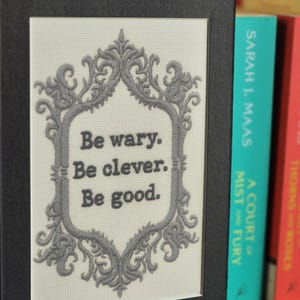 Op de afbeelding: Een ingelijst kunstwerk met de tekst "Be wary. Be clever. Be good." in een decoratieve grijze lijst. Het kunstwerk staat tegen een neutrale achtergrond en is zwart omlijst. Boeken zijn zichtbaar op de achtergrond.