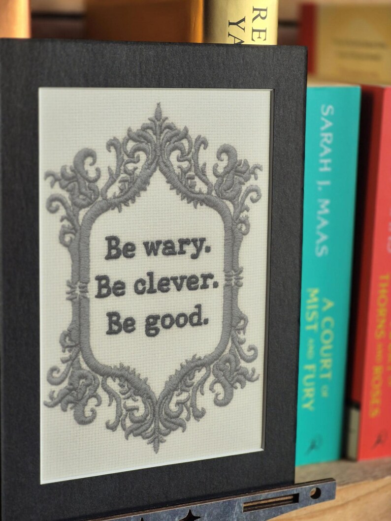 Op de afbeelding: Een ingelijst kunstwerk met de tekst "Be wary. Be clever. Be good." in een decoratieve grijze lijst. Het kunstwerk staat tegen een neutrale achtergrond en is zwart omlijst. Boeken zijn zichtbaar op de achtergrond.