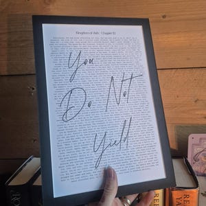 May include: A framed print with the text "You Do Not Yield" in a handwritten style, overlaying printed text. The print is in a black frame, held by a person. The background includes books and wooden paneling.