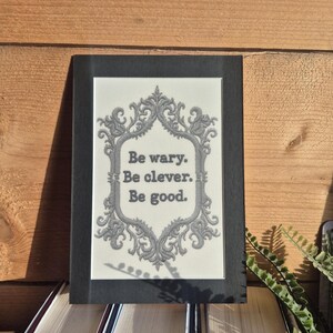Op de afbeelding: Een ingelijste print met de tekst "Be wary. Be clever. Be good." De tekst staat in een decoratief lettertype tegen een cr&egrave;mekleurige achtergrond en is omlijst door een sierlijke grijze rand. De lijst is zwart en de print ligt op een stapel boeken.