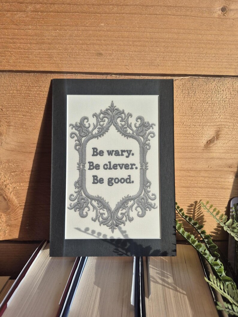 Op de afbeelding: Een ingelijste print met de tekst "Be wary. Be clever. Be good." De tekst staat in een decoratief lettertype tegen een cr&egrave;mekleurige achtergrond en is omlijst door een sierlijke grijze rand. De lijst is zwart en de print ligt op een stapel boeken.