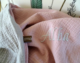 Puede incluir: Una manta de muselina de doble capa con un lado rosa empolvado y otro gris claro. El lado rosa está bordado con el nombre Alba en letra cursiva blanca. Una pequeña etiqueta de tela marrón con texto en blanco está unida al borde.