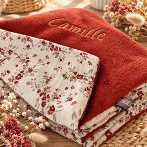 Peut inclure: Une couverture pliée présentant une face unie de couleur rouille avec le prénom Camille brodé en écriture cursive et une face crème ornée d'un petit motif floral rouge et marron. Une petite étiquette en tissu est visible sur le bord.