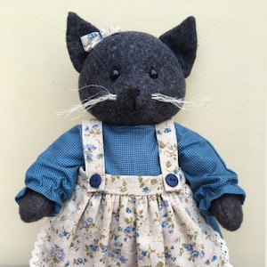 Peut inclure: Une poupée chat en peluche faite à la main avec une tête, des oreilles et des pattes gris foncé. Elle porte une robe en vichy bleu avec un tablier à imprimé floral et un nœud assorti. Le tablier a une bordure en dentelle blanche. Le chat a des moustaches blanches et des yeux en bouton.