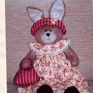 May include: A handmade plush bunny doll with a brown body, large ears, and a pink hat. The bunny wears a floral dress with a white lace collar and holds a pink striped purse. The doll's face is stitched with black thread.