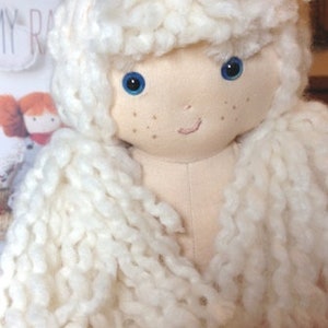 May include: A handmade doll with long, white yarn hair, blue eyes, and a light pink complexion. The doll has a simple smile and is made of soft fabric.