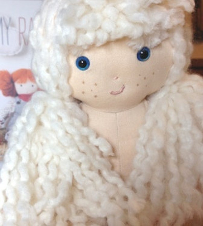 May include: A handmade doll with long, white yarn hair, blue eyes, and a light pink complexion. The doll has a simple smile and is made of soft fabric.