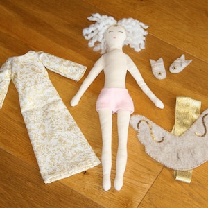May include: A handmade doll with white yarn hair, a pink skirt, and a gold and white dress. The doll is wearing a pair of white felt shoes and a beige felt angel wing accessory.