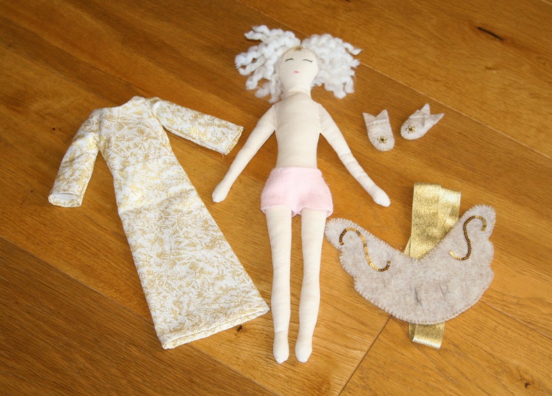 May include: A handmade doll with white yarn hair, a pink skirt, and a gold and white dress. The doll is wearing a pair of white felt shoes and a beige felt angel wing accessory.