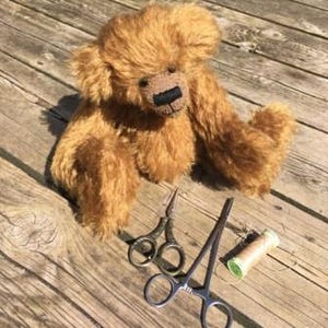 May include: A handcrafted, golden-brown teddy bear with a stitched black nose and a fluffy texture. Next to the bear are small silver scissors, a silver clamp, and a reel of thread, all on a wooden surface.
