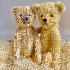 May include: Two vintage-style teddy bears with light brown fur. The bears have black stitched noses and expressive eyes. They are sitting in a bed of shredded paper, creating a cozy feel. These charming bears would be a lovely addition to any collection.