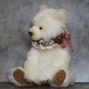 Op de afbeelding: Een handgemaakte, crèmekleurige teddybeer met een gedetailleerd ontwerp. De beer heeft een bruine neus en poten en draagt een bloemvormige kraag met een kleine bel en een roze lint. De beer zit.