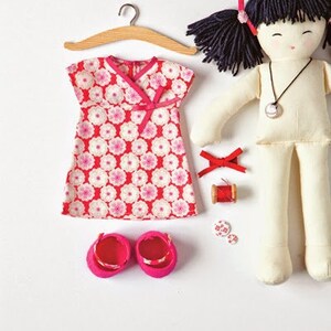 May include: A handmade doll with black hair and a white dress. The doll is wearing a pink and white floral dress with a pink bow. The doll is also wearing pink shoes. The doll is sitting on a white surface.