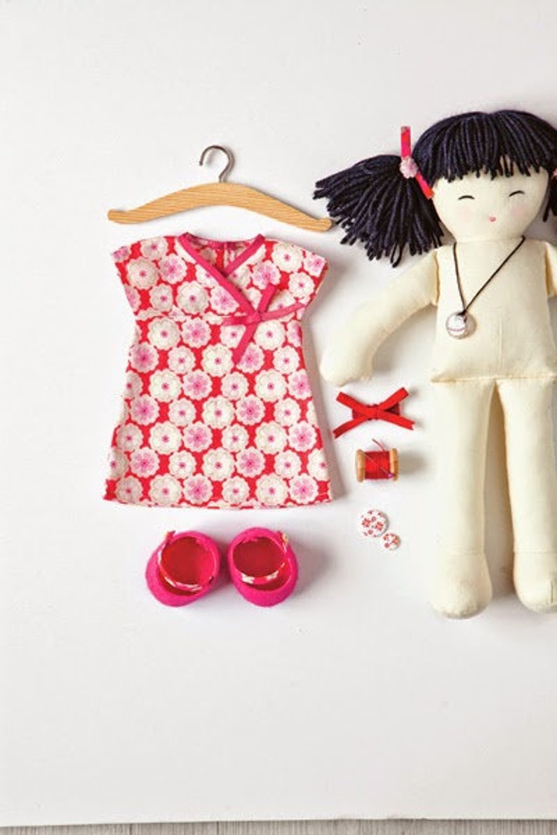 May include: A handmade doll with black hair and a white dress. The doll is wearing a pink and white floral dress with a pink bow. The doll is also wearing pink shoes. The doll is sitting on a white surface.
