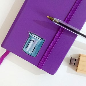 May include: A purple notebook with a pink ribbon bookmark and a beaker sticker. A silver pen and a wooden USB drive are also on the white surface.