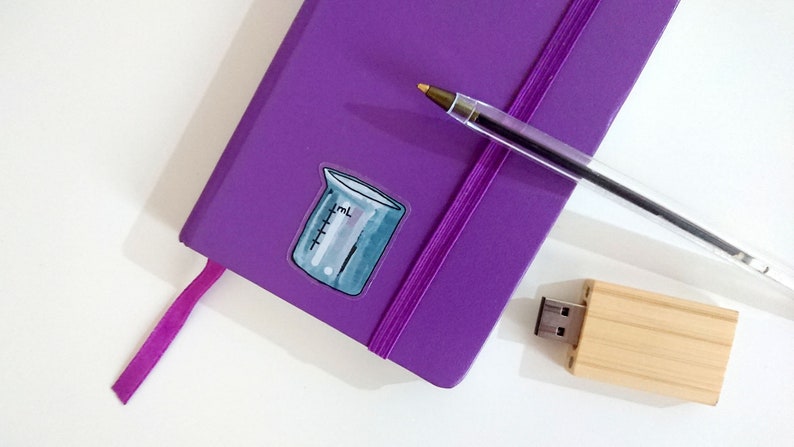 May include: A purple notebook with a pink ribbon bookmark and a beaker sticker. A silver pen and a wooden USB drive are also on the white surface.