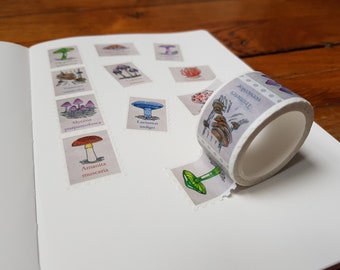 May include: A sheet of decorative washi tape with mushroom illustrations and names. The tape is white with a light gray background and features different types of mushrooms, including Amanita muscaria, Mycena purpureofusca, and Lactarius indigo. The tape is partially unrolled and shows the design.