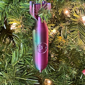 May include: A iridescent purple and green bomb-shaped Christmas ornament with a silver circle on the side. The ornament is hanging on a green artificial Christmas tree.
