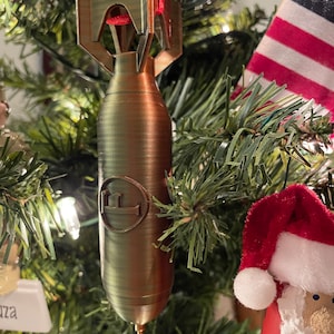 May include: A bronze-colored Christmas tree ornament shaped like a bomb, hanging from a red ribbon. The ornament has a detailed design with a circular emblem and is suspended among green pine needles and Christmas lights.