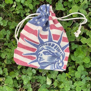 May include: A small drawstring bag made of fabric with a red, white, and blue American flag design. The bag features a blue silhouette of the Statue of Liberty.