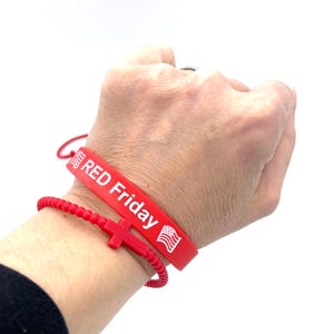 May include: Red silicone bracelet with the text "RED Friday" and an American flag. A red beaded bracelet with a red cross charm is worn on top of the silicone bracelet.
