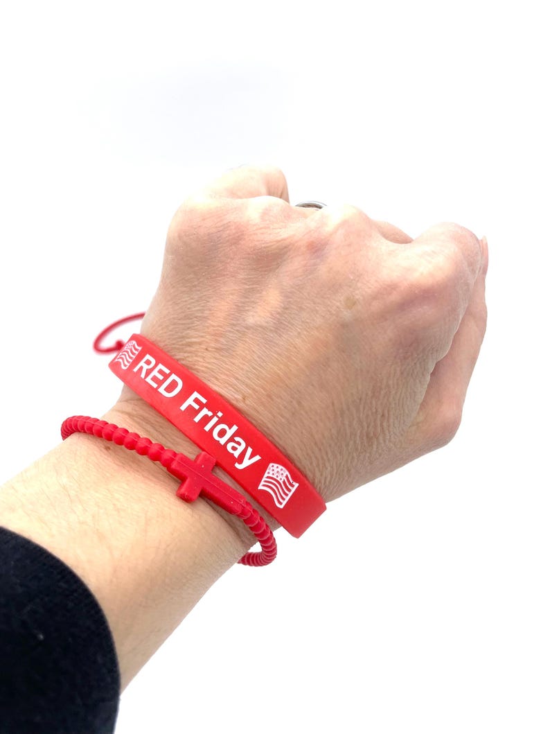 May include: Red silicone bracelet with the text "RED Friday" and an American flag. A red beaded bracelet with a red cross charm is worn on top of the silicone bracelet.