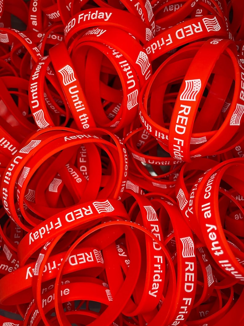 May include: A pile of red silicone wristbands with the text "RED Friday" and "Until they all come home" printed in white. The wristbands also feature a small American flag graphic.