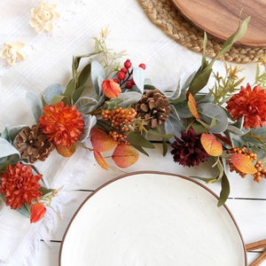 May include: A decorative autumn garland featuring artificial orange and dark red chrysanthemum flowers, brown pinecones, red berries, orange berry clusters, and sage green lamb's ear leaves arranged on a flexible vine base for seasonal display.