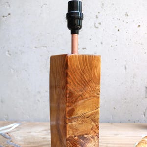 May include: A handcrafted wooden lamp with a natural wood grain finish. The lamp features a black lamp socket, a copper pipe, and a rectangular wooden base. The lamp is on a wooden surface.