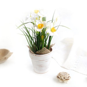 May include: A white ceramic pot with a burlap base holds a bouquet of white daffodils with yellow centers. The pot is sitting on a white surface.