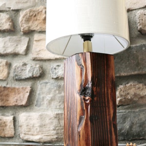 May include: A rustic wooden table lamp with a cream-colored lampshade. The lamp base is a dark brown, rectangular block of wood with visible grain and knots. The lamp is set against a stone wall, with pine cones at the base.