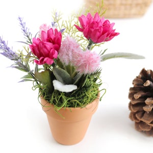 May include: A small terracotta pot filled with green moss and artificial flowers. The flowers are pink, purple, and white. The pot is sitting on a white surface.