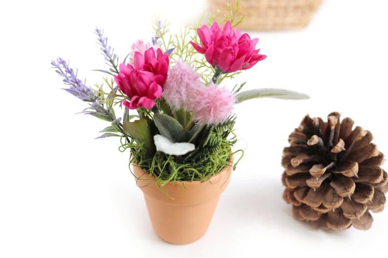 May include: A small terracotta pot filled with green moss and artificial flowers. The flowers are pink, purple, and white. The pot is sitting on a white surface.
