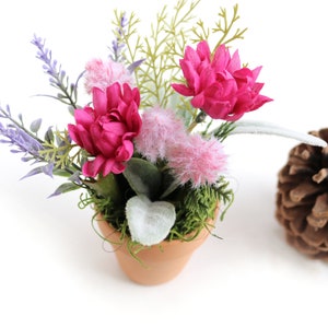 May include: A small terracotta pot filled with a floral arrangement featuring pink and purple flowers, green foliage, and white fuzzy accents. The pot is sitting on a white surface.